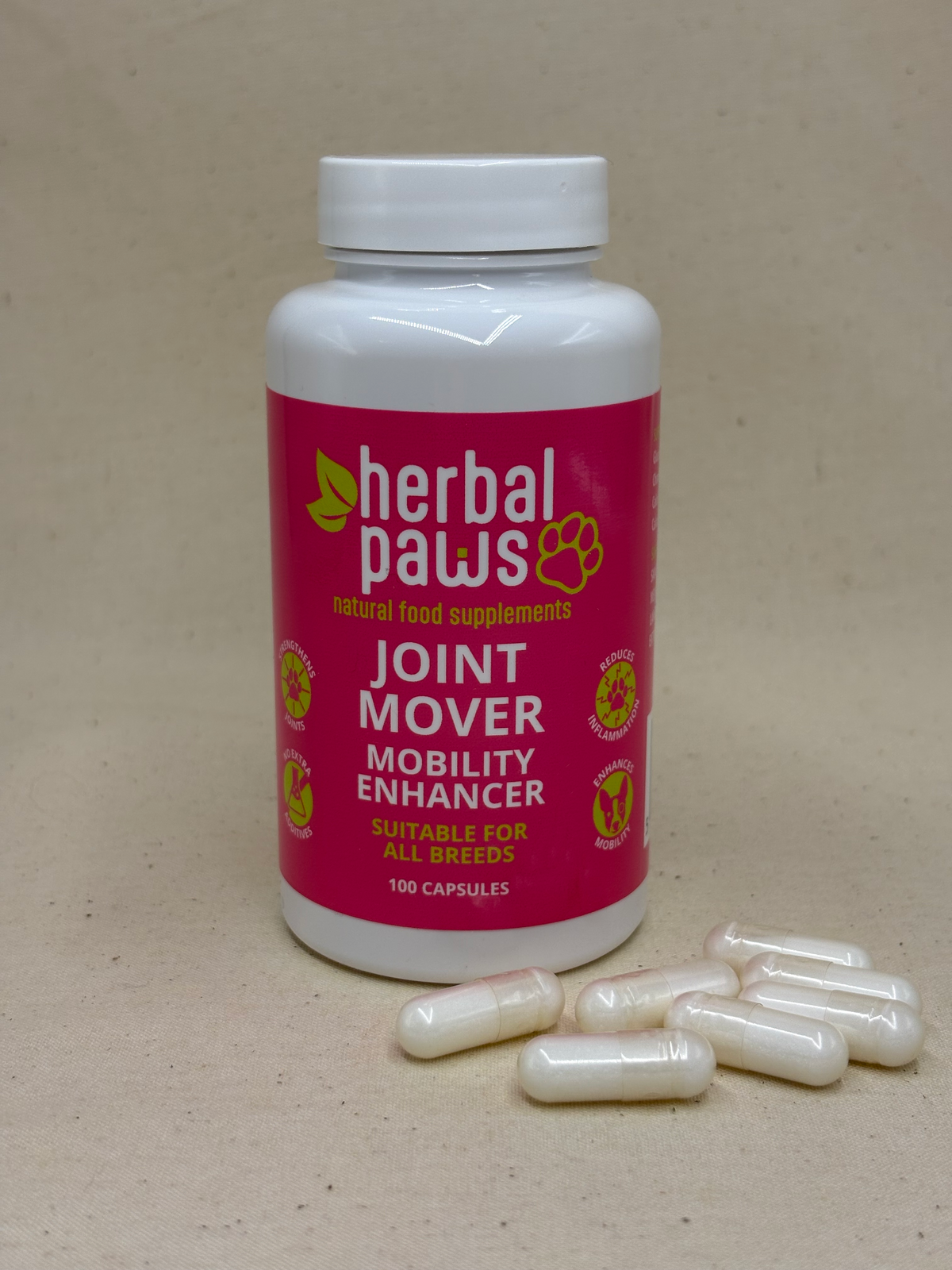 A white bottle of Herbal Paws Joint Mover Mobility Enhancer with a bright pink label, shown on a beige background with several white capsules scattered in front. Natural supplement for dogs